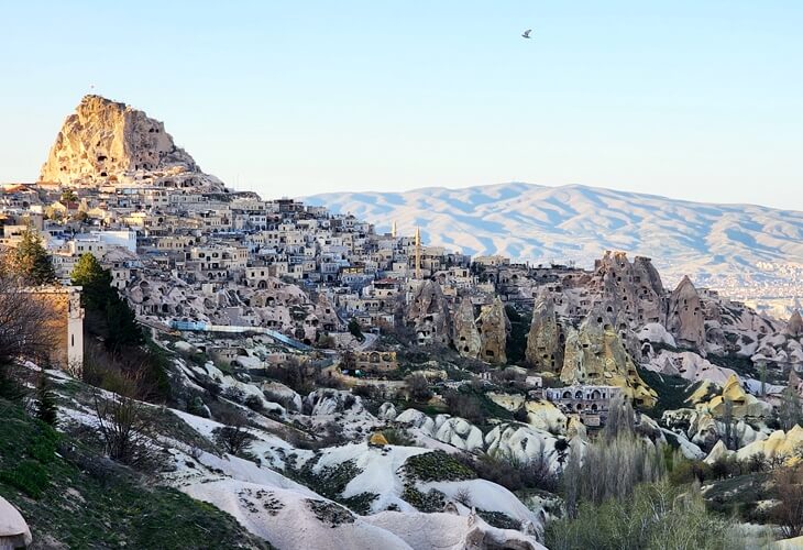 Uçhisar Kalesi ve çevresindeki kaya evlerle Kapadokya’nın genel manzarası, dağ silüeti eşliğinde